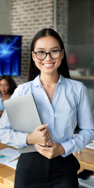 Portrait Of Smiling Asian Female Employee Posing In Office Interior During Meeting With Colleagues, Young Korean Business Lady Holding Laptop, Ready For Presentation In Board Room, Closeup